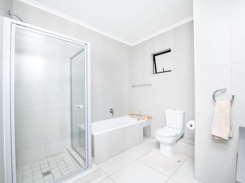 A modern, neutral-toned bathroom featuring a glass-enclosed shower, a built-in bathtub, a toilet, and tiled flooring throughout.