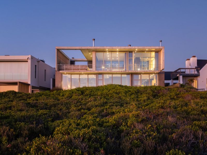 Modern multi-level residence featuring extensive glass facades, concrete architectural elements, and a spacious upper-level balcony overlooking the coastal landscape.