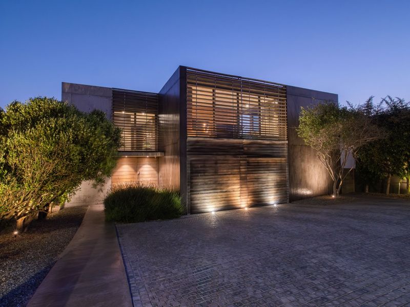 Modern architectural residence featuring a concrete and timber facade, integrated louvre shutters, and a paved cobblestone driveway with ambient exterior lighting.
