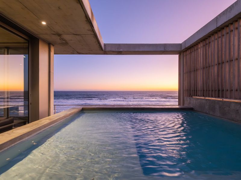 A modern, high-end pool area featuring exposed concrete architecture and a timber privacy screen, offering stunning, unobstructed ocean views at sunset.