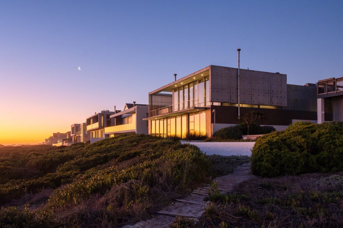 A modern architectural home featuring concrete construction and expansive glass windows, set against a coastal landscape at sunset.