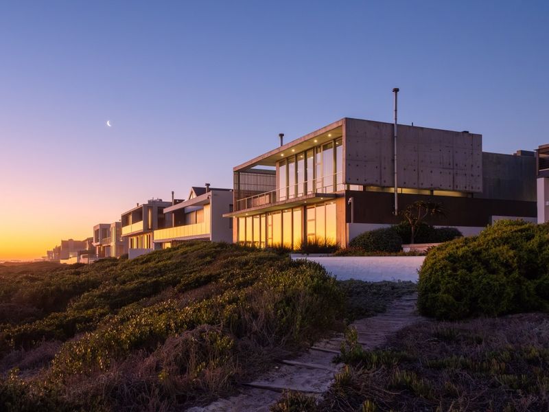 A modern architectural home featuring concrete construction and expansive glass windows, set against a coastal landscape at sunset.