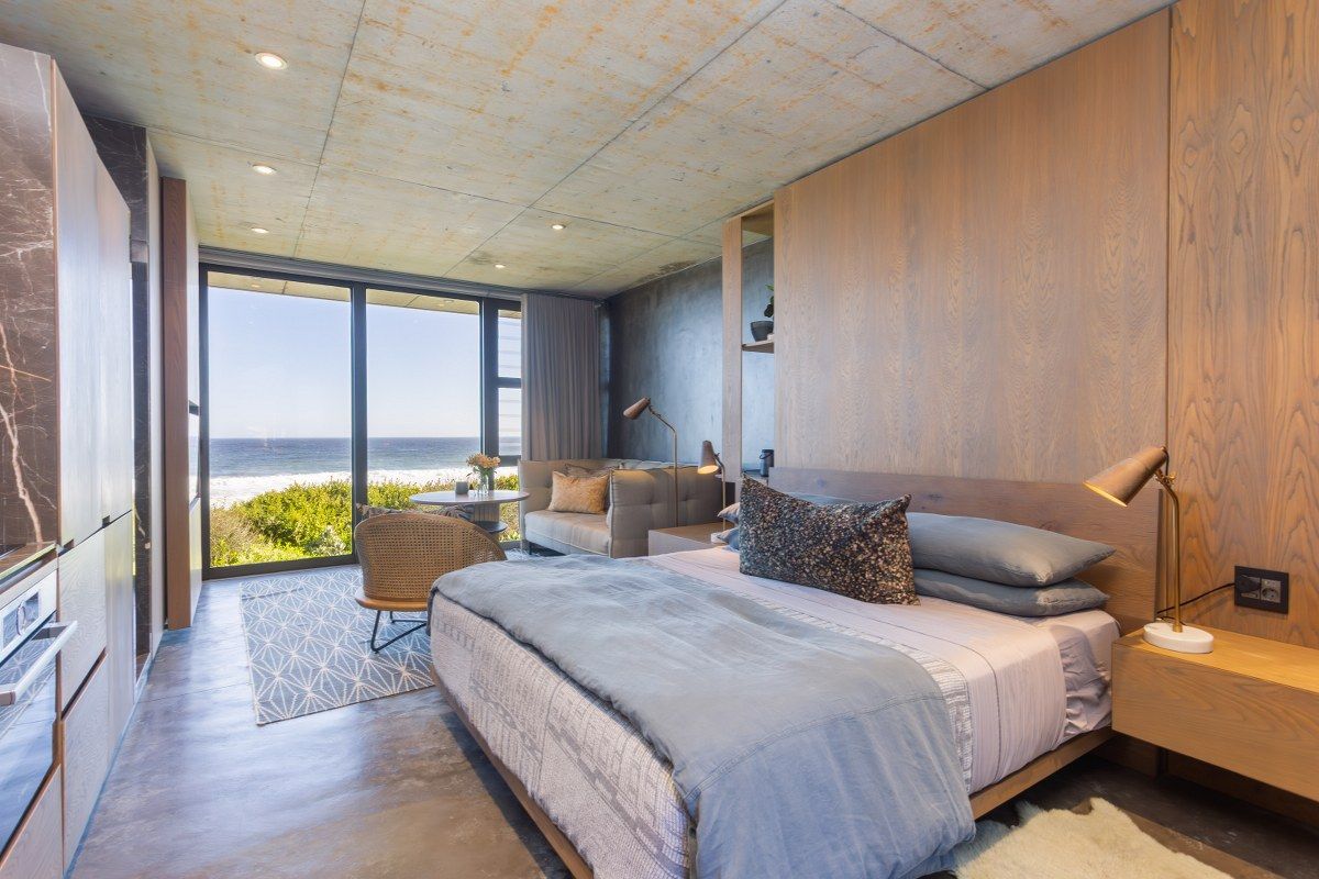 Modern bedroom featuring polished concrete floors, exposed concrete ceiling, timber wall paneling, and floor-to-ceiling glass doors with direct ocean views.
