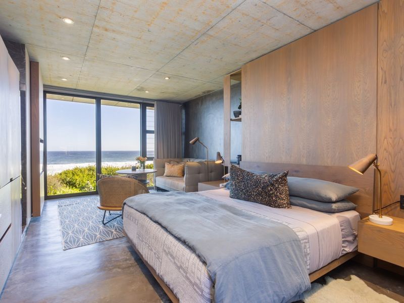 Modern bedroom featuring polished concrete floors, exposed concrete ceiling, timber wall paneling, and floor-to-ceiling glass doors with direct ocean views.