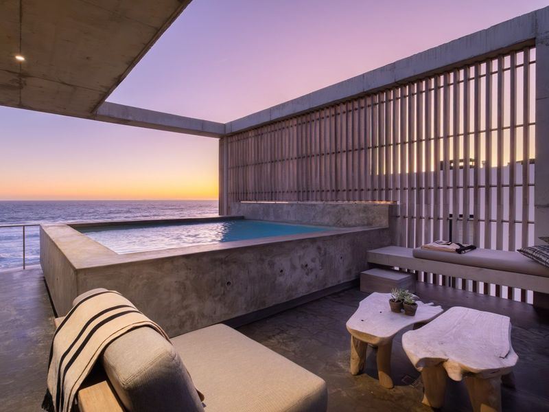 A modern outdoor terrace featuring a concrete plunge pool, built-in bench seating, and timber privacy screens with panoramic ocean views at sunset.