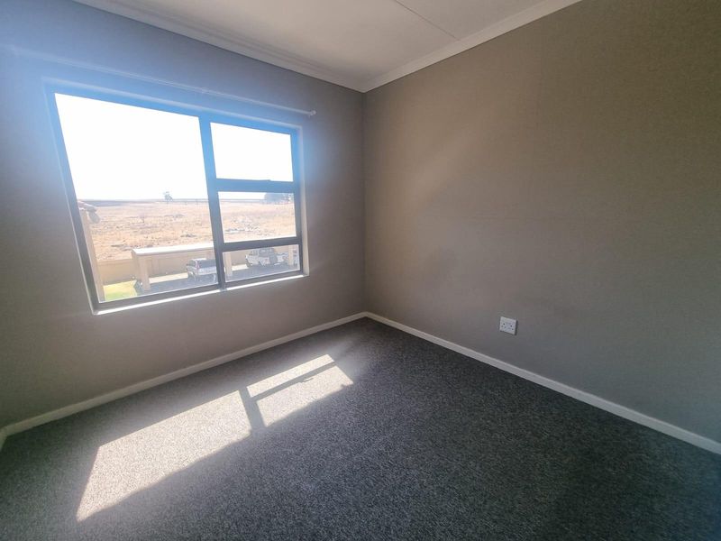 A bright, empty bedroom featuring neutral grey walls, carpeted flooring, and a large window with views of the surrounding area.