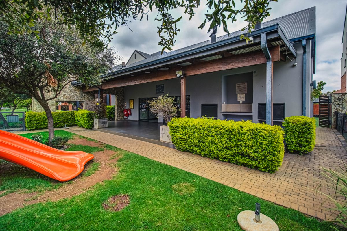 A well-maintained rear exterior area featuring a covered patio with a built-in braai, stone accents, and a paved walkway leading to a garden space.