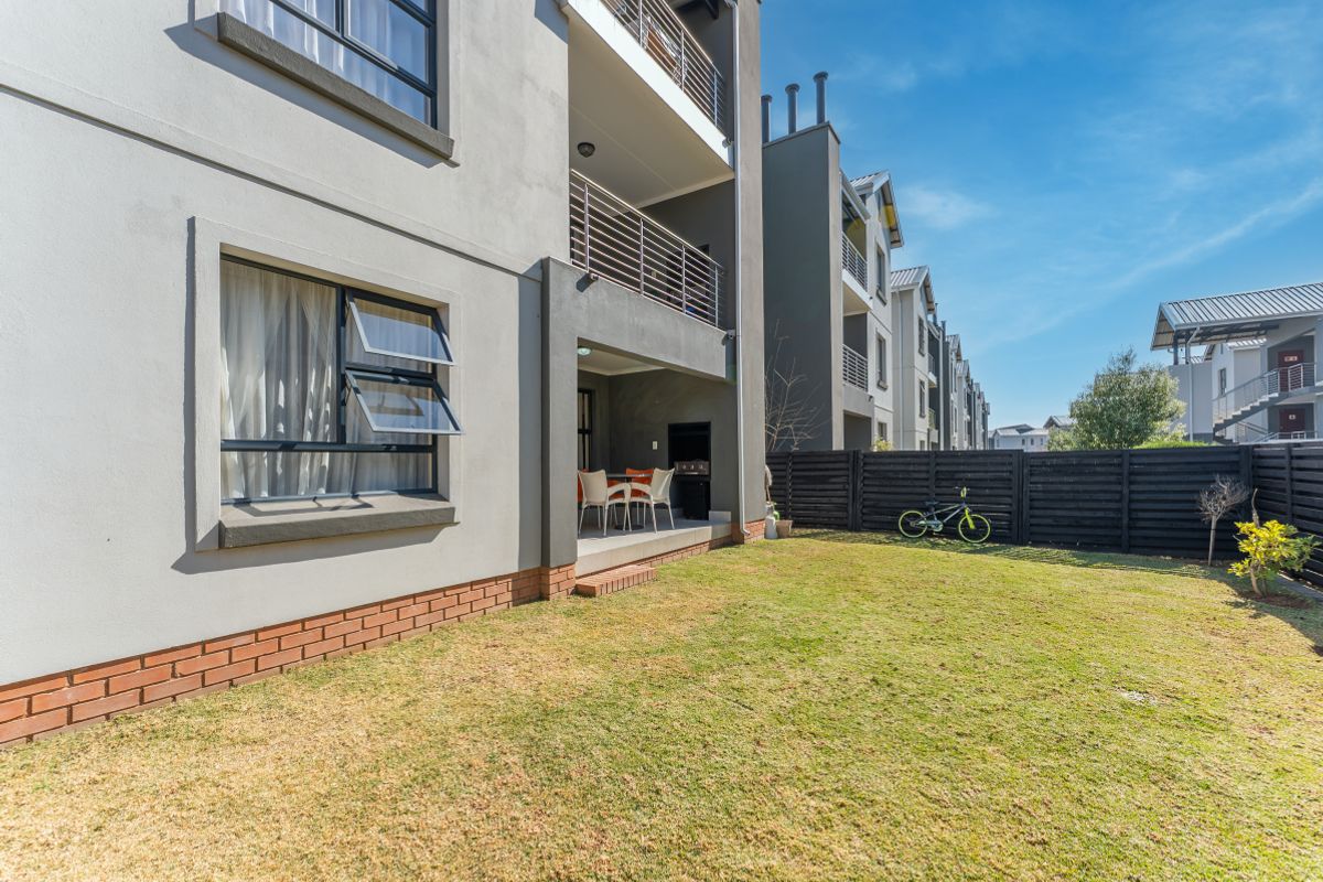 Rear view of the apartment building featuring a covered patio with a built-in braai, overlooking a private garden area with timber fencing.