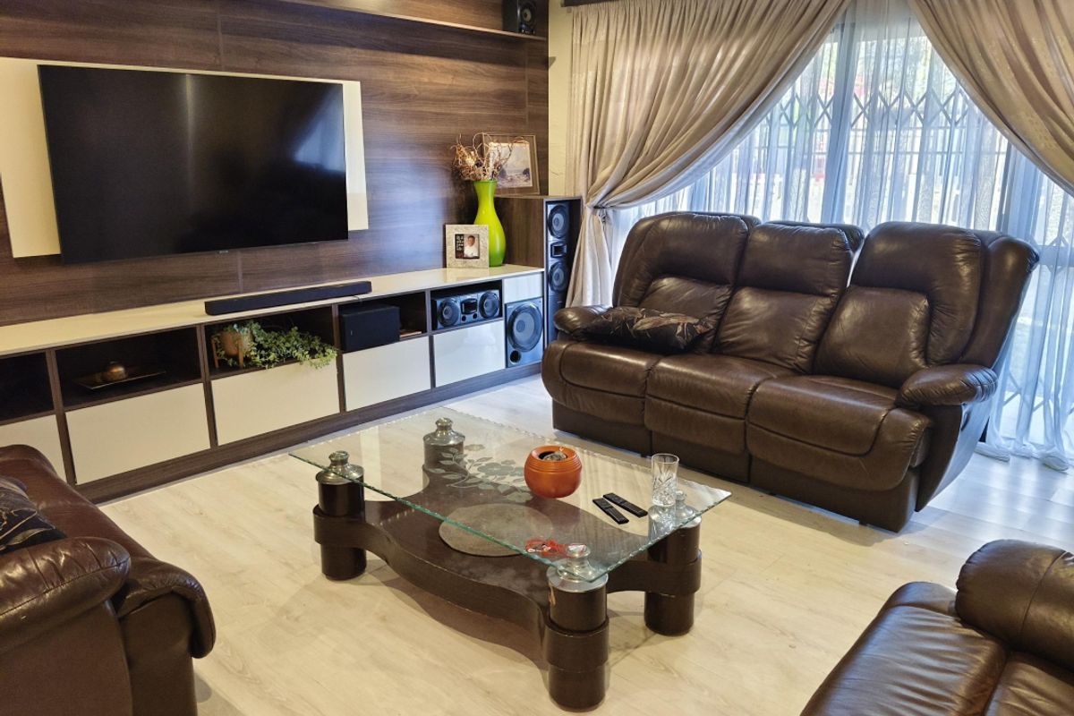 A well-maintained living room featuring a custom built-in media wall unit, light-toned laminate flooring, and large windows with integrated security gates.