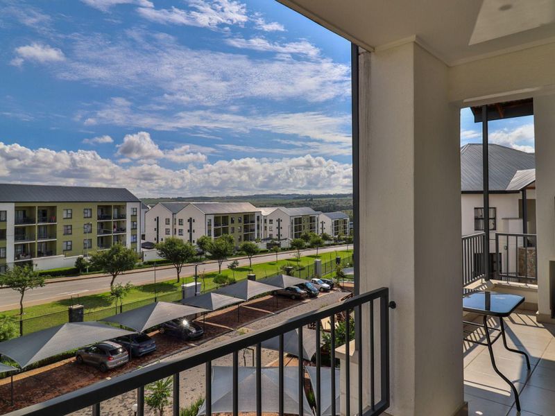 A covered balcony featuring tiled flooring, a metal balustrade, and a built-in braai, offering an elevated view of the surrounding apartment complex and parking area.