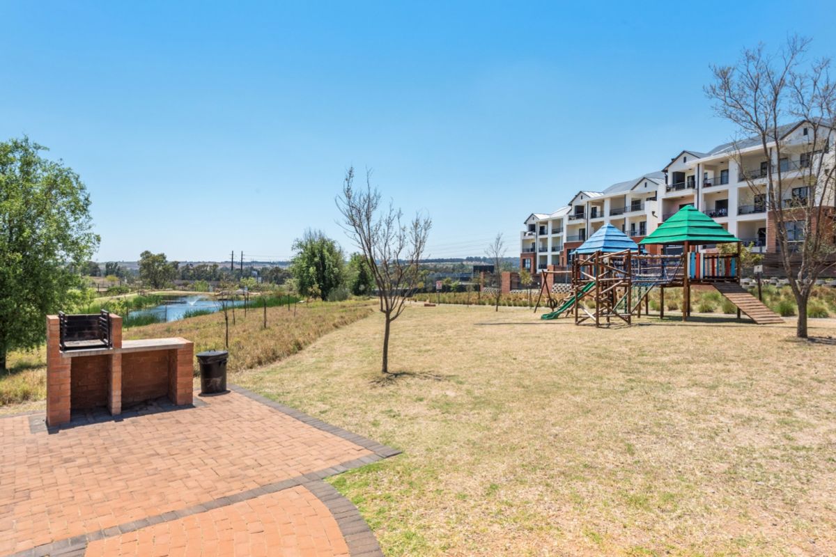 Communal outdoor area featuring a paved patio with a built-in brick braai, children's playground equipment, and views over the landscaped garden and pond.