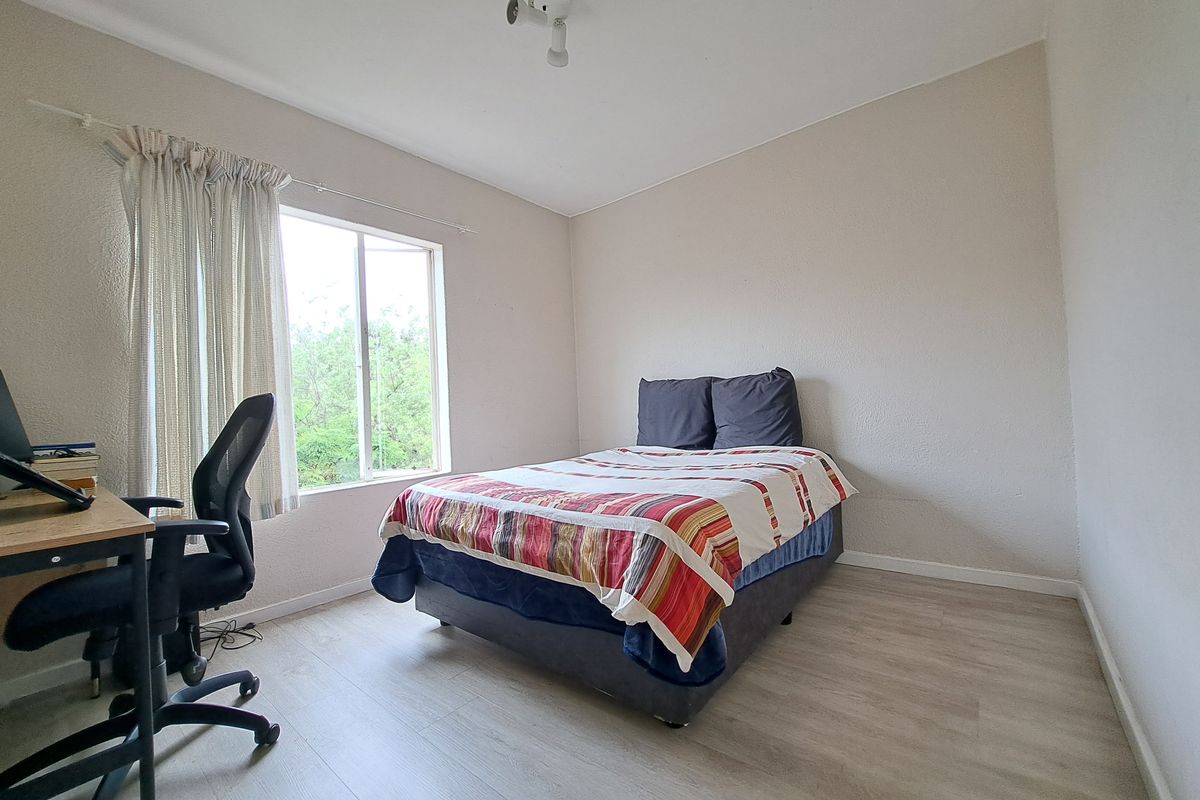 A functional bedroom featuring light-toned laminate flooring and a large window that provides natural light and a view of the surrounding greenery.