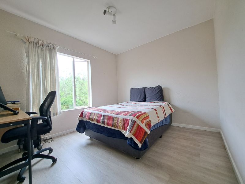 A functional bedroom featuring light-toned laminate flooring and a large window that provides natural light and a view of the surrounding greenery.