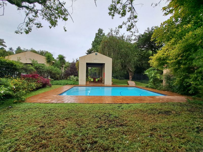 A well-maintained swimming pool area featuring a brick surround and a covered gazebo, set within a lush, landscaped garden environment.