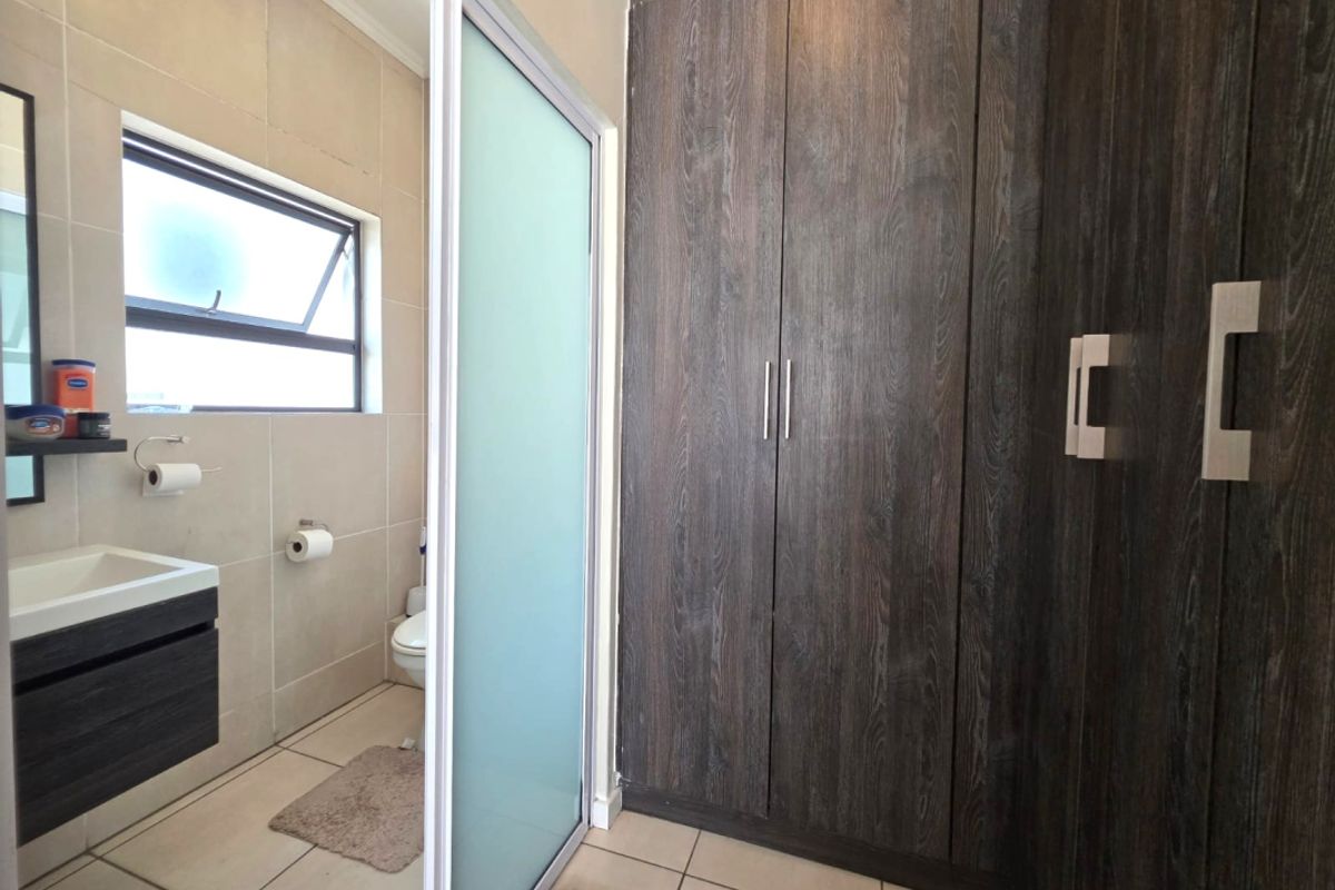 Modern bathroom featuring dark wood-finish built-in cupboards, a sleek vanity unit, and neutral wall and floor tiling with a frosted glass sliding door.