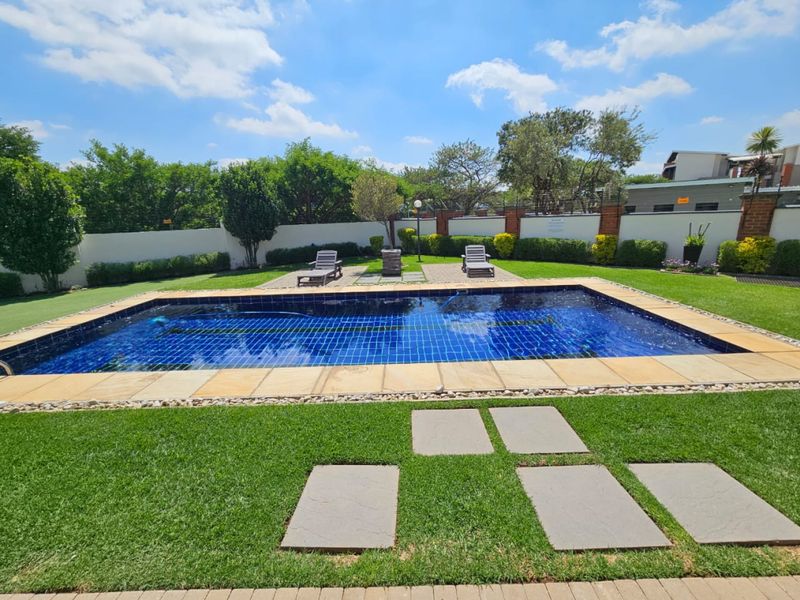 A well-maintained outdoor swimming pool area featuring a tiled pool, stone paving, and landscaped lawn surrounded by a perimeter wall.