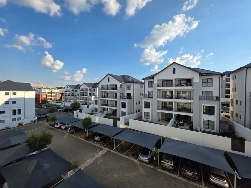 An aerial view of a modern apartment complex featuring multi-story residential buildings, covered parking bays, and paved communal areas under a clear sky.