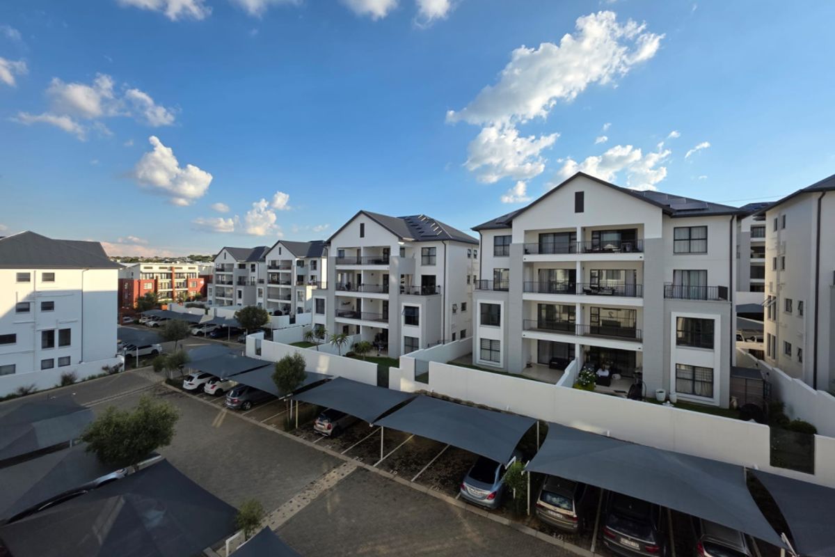 An aerial view of a modern apartment complex featuring multi-story residential buildings, covered parking bays, and paved communal areas under a clear sky.