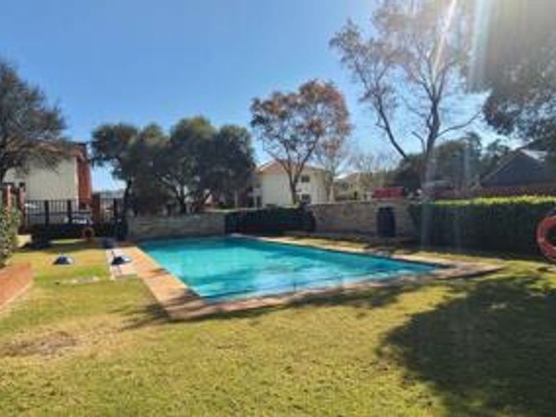A well-maintained communal swimming pool area featuring a clear pool, stone boundary walls, and surrounding landscaped lawn and hedging.