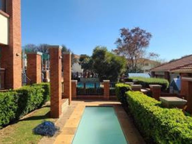 Outdoor swimming pool area featuring brick pillars, paved decking, and manicured hedge borders within a residential apartment complex.