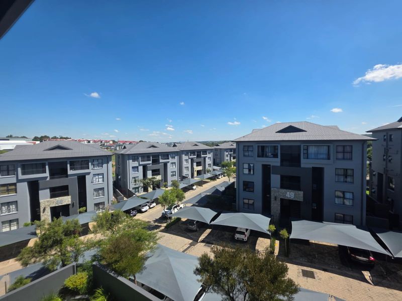 An elevated aerial view of a modern apartment complex featuring multiple grey multi-story buildings, paved driveways, and covered shade cloth parking bays.