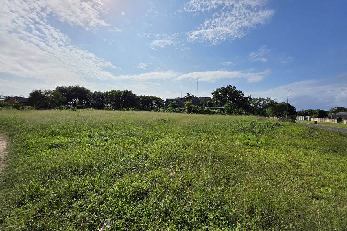 A wide-angle view of a vacant residential plot featuring open grassland and surrounding mature trees under a clear blue sky.