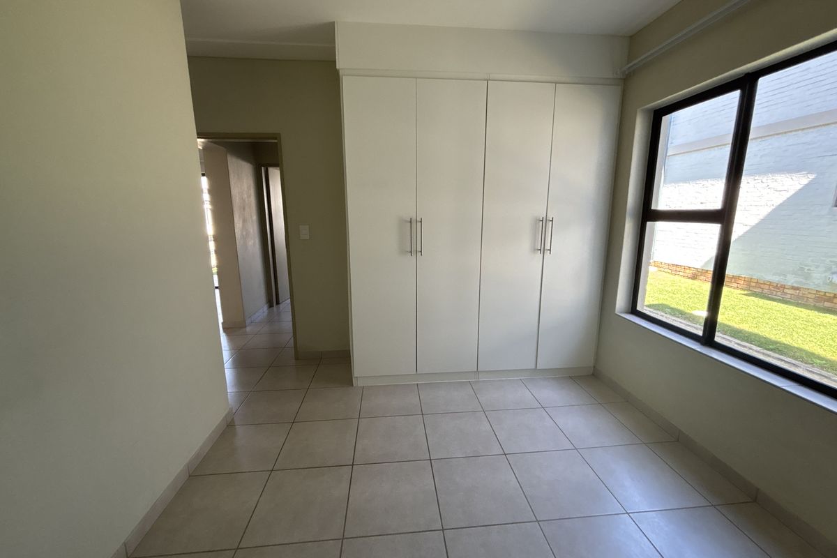 A bright, empty bedroom featuring neutral tiled flooring, built-in cupboards, and a large window overlooking the garden area.
