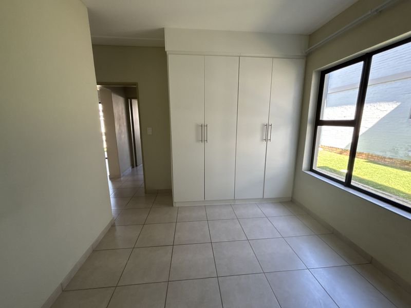 A bright, empty bedroom featuring neutral tiled flooring, built-in cupboards, and a large window overlooking the garden area.