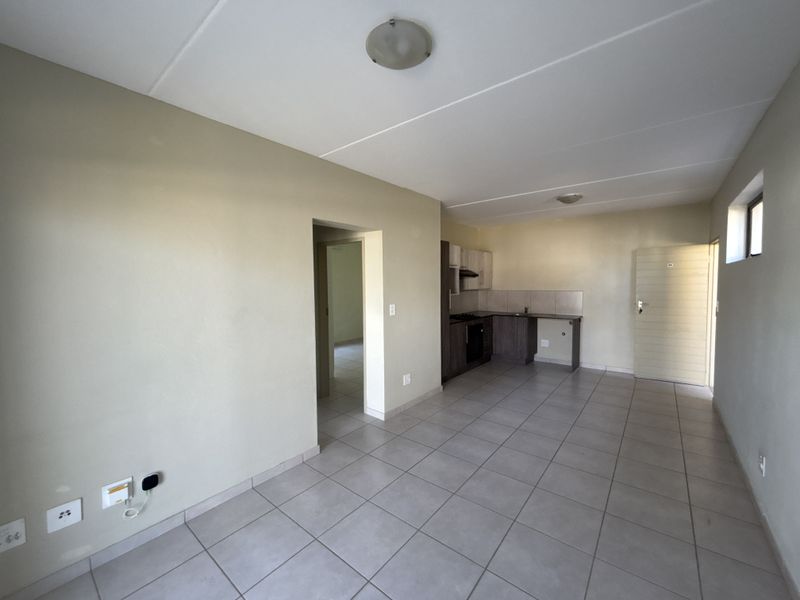 An open-plan living and kitchen area featuring neutral tiled flooring, built-in cabinetry, and integrated kitchen appliances in a clean, vacant space.