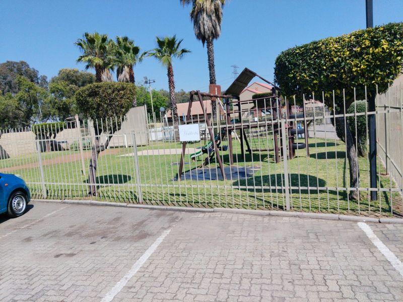 A communal outdoor area featuring a children's playground, landscaped garden, and steel perimeter fencing, adjacent to a paved parking lot.