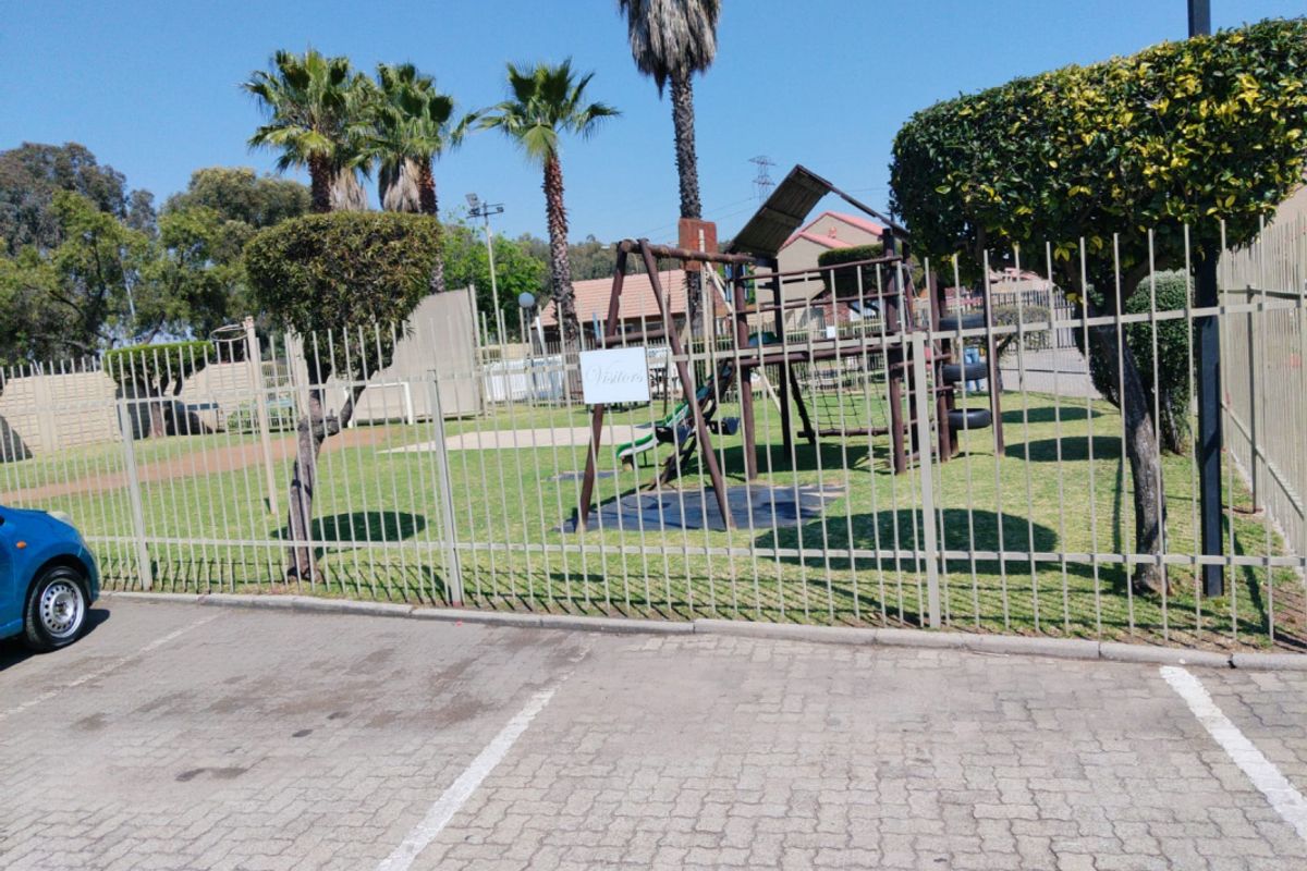 A communal outdoor area featuring a children's playground, landscaped garden, and steel perimeter fencing, adjacent to a paved parking lot.