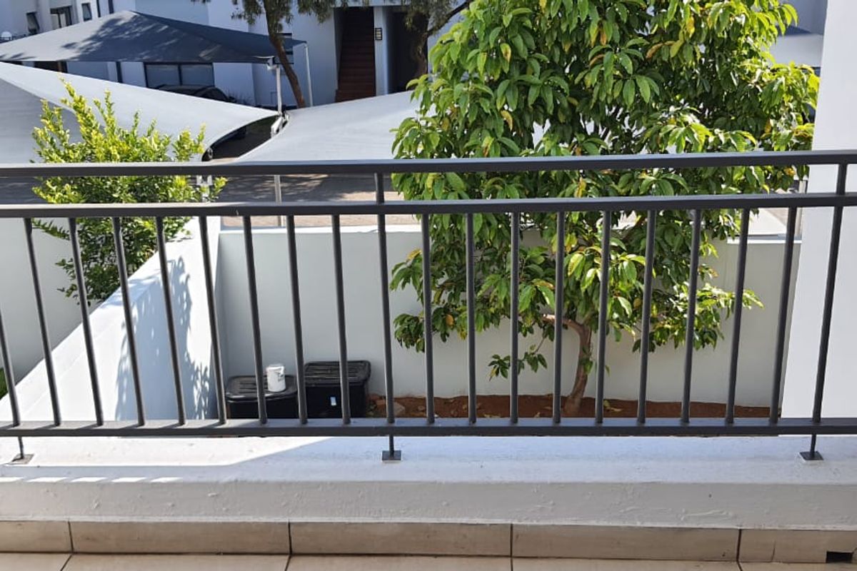 A private balcony featuring a metal balustrade and tiled flooring, overlooking the complex grounds and parking area.