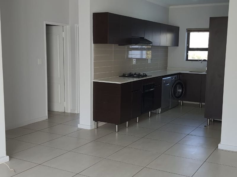Modern open-plan kitchen featuring dark cabinetry, gas hob, under-counter oven, extractor fan, and neutral tiled flooring in a clean, vacant space.