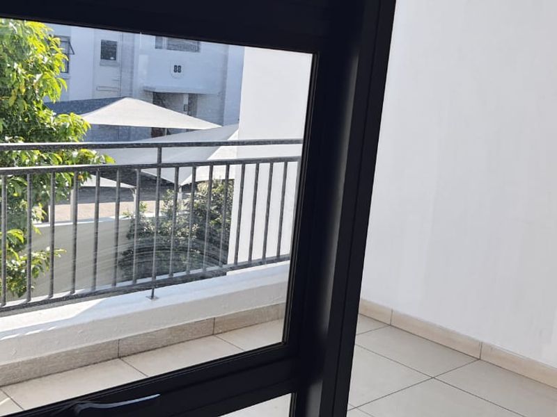 A clean, tiled balcony featuring a metal balustrade and sliding glass door access, overlooking the complex grounds.