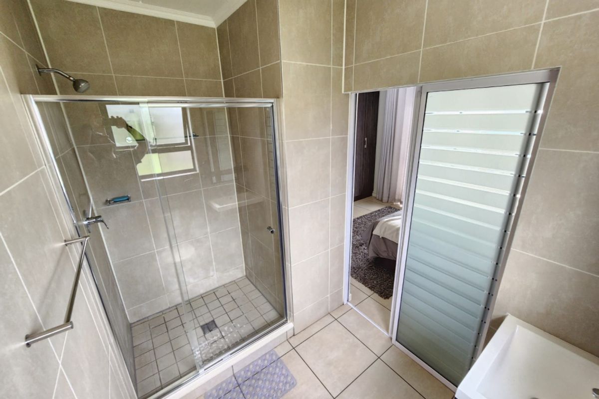 A modern bathroom featuring a glass-enclosed shower, neutral wall and floor tiling, and a vanity basin, with direct access to the adjacent bedroom.