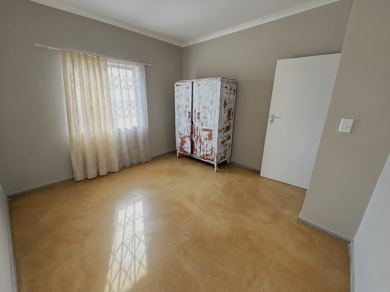A clean, neutral-toned bedroom featuring polished concrete flooring, a security gate on the window, and a simple internal door.