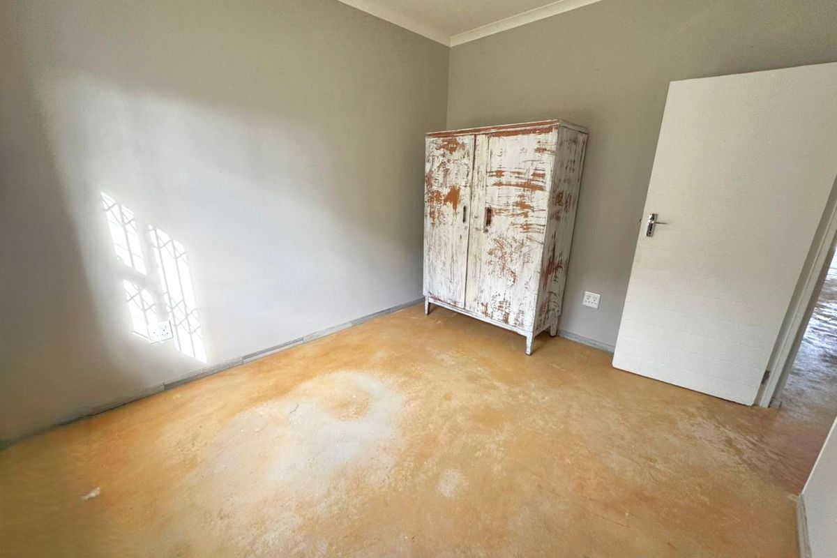 A simple bedroom featuring neutral walls, a security gate on the window, and exposed concrete flooring.