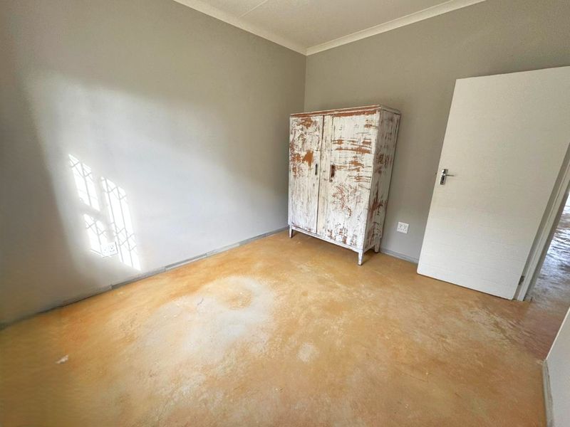 A simple bedroom featuring neutral walls, a security gate on the window, and exposed concrete flooring.