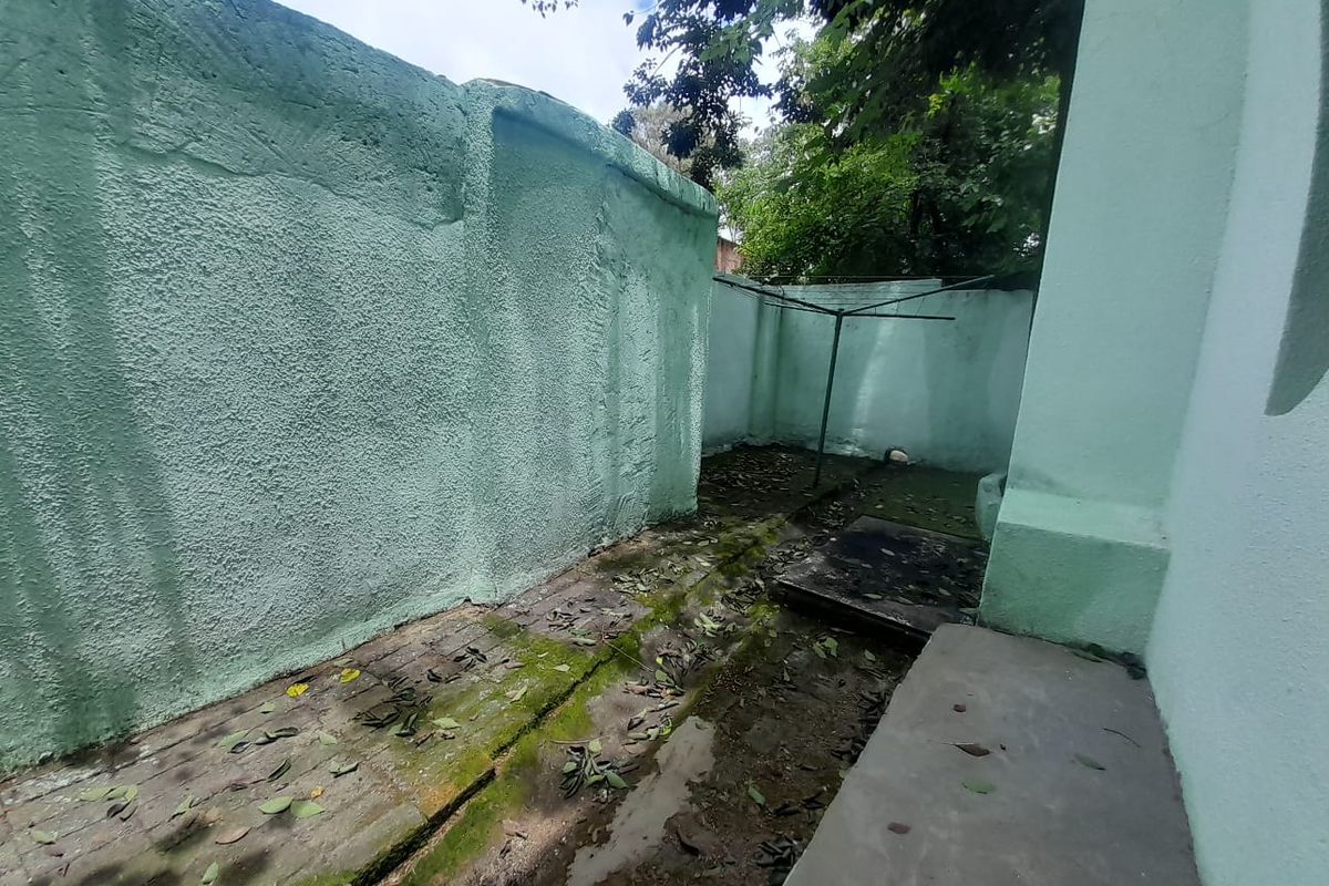 A narrow, paved rear exterior area featuring boundary walls and drainage channels, currently showing signs of weathering and moss accumulation.