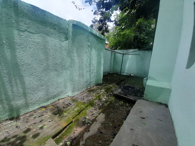 A narrow, paved rear exterior area featuring boundary walls and drainage channels, currently showing signs of weathering and moss accumulation.