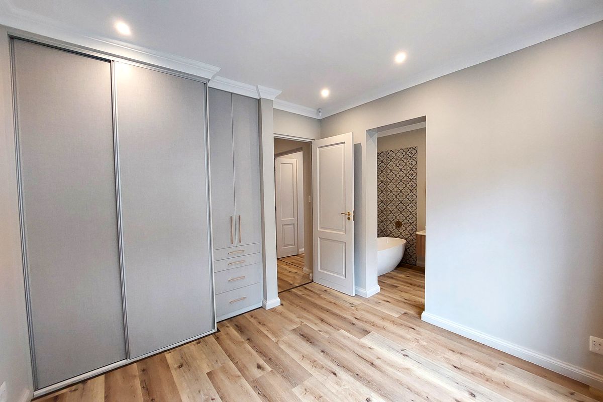 A bright, modern bedroom featuring light wood-look laminate flooring, extensive built-in cabinetry, and direct access to an en-suite bathroom.