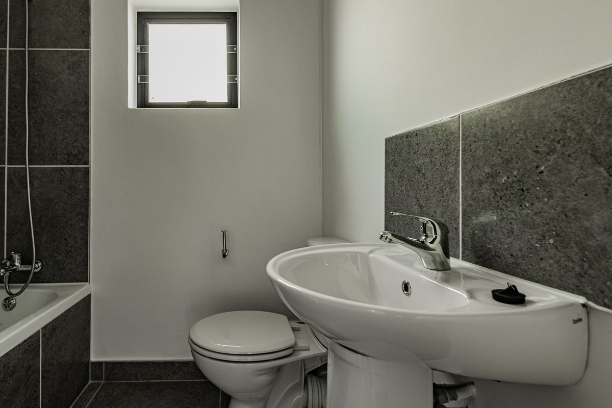 A clean, modern bathroom featuring a pedestal basin, chrome fixtures, a bathtub with shower mixer, and neutral grey floor and wall tiling.