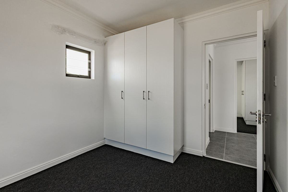A clean, empty bedroom featuring dark carpeting, a built-in white wardrobe, and a small louver window, leading into a tiled hallway.