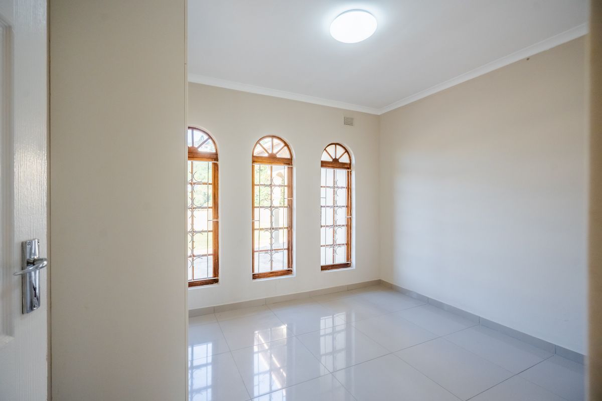 A bright, empty bedroom featuring polished tiled floors and three distinctive arched wooden windows with security bars.