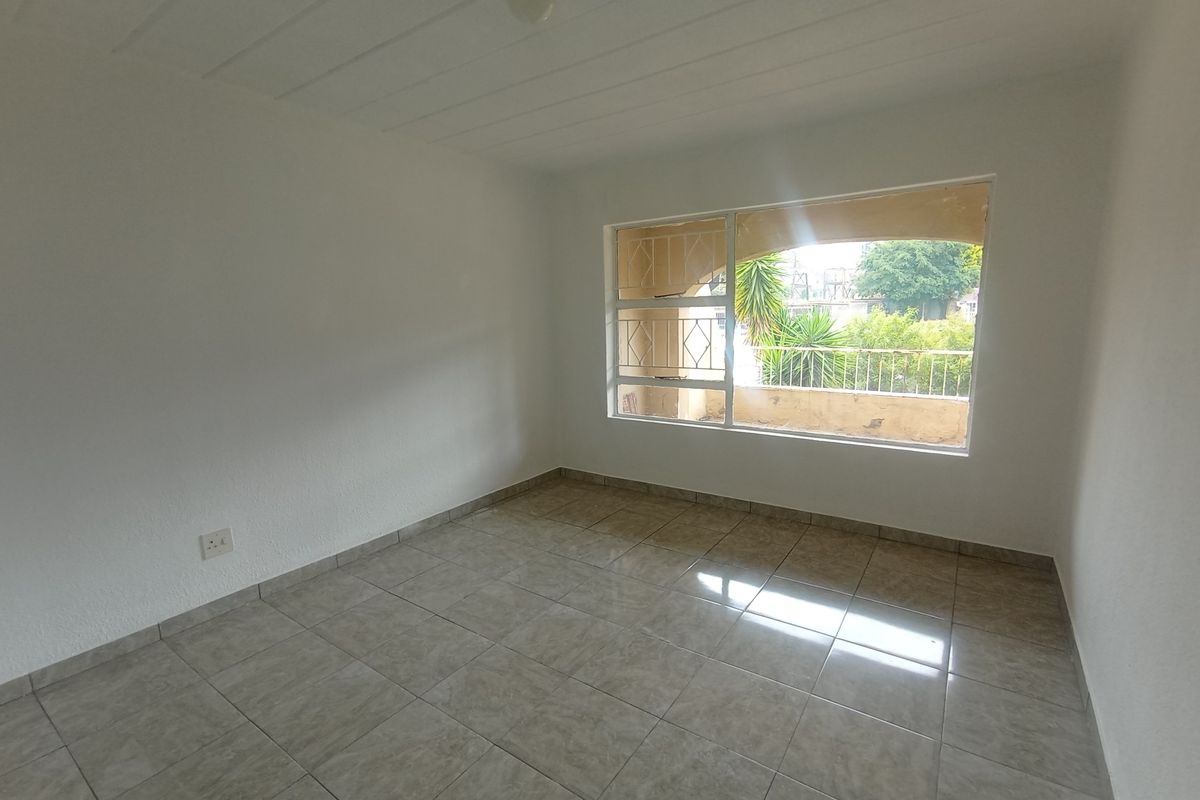 A bright, empty bedroom featuring neutral tiled flooring, a large window with security bars, and a view of the exterior balcony.
