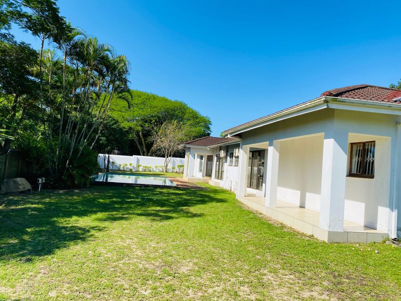 Rear view of the property featuring a covered patio, a swimming pool, and a spacious garden area with established trees and perimeter walling.