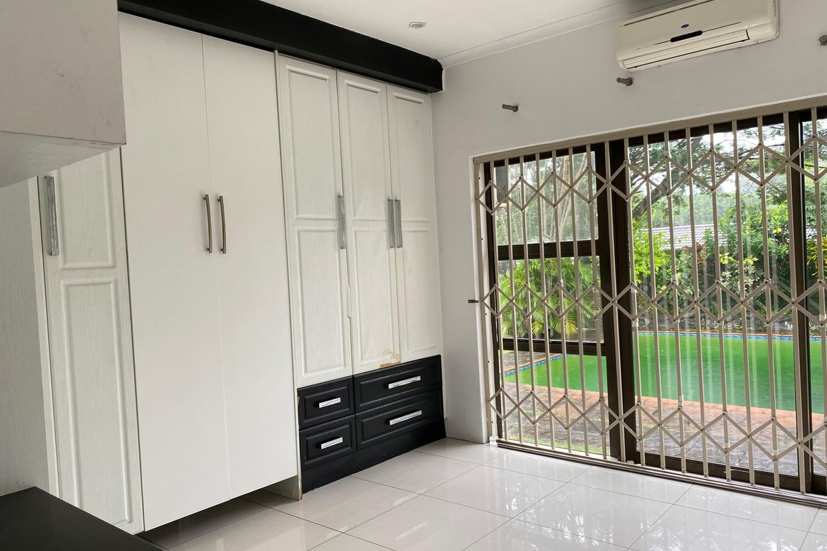 A bright bedroom featuring built-in cupboards, tiled flooring, and a sliding glass door with a security gate, offering direct access to the pool area.
