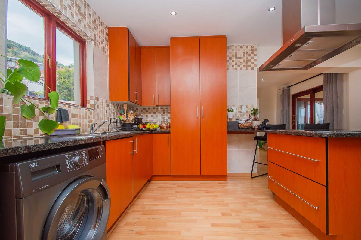 A functional kitchen featuring orange-toned cabinetry, granite countertops, a stainless steel extractor fan, and integrated space for a washing machine.