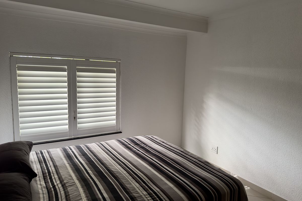 A bright bedroom featuring white walls, tiled flooring, and fitted plantation shutters on the window.