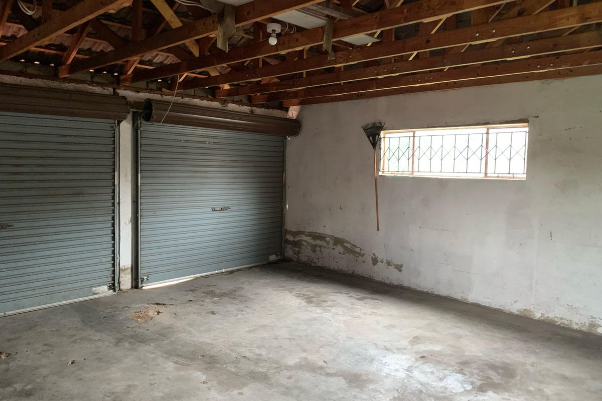 A spacious double garage featuring exposed timber roof trusses, concrete flooring, and dual roller shutter doors, requiring maintenance and cosmetic updates.